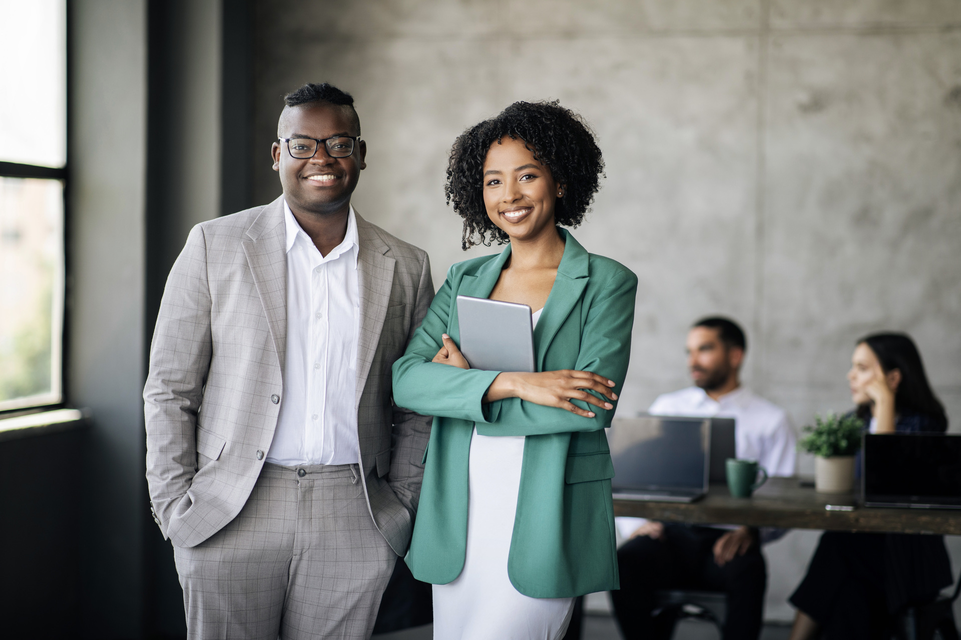 A smiling man in a light gray plaid suit and a smiling woman in a green blazer holding a tablet stand together in a modern office